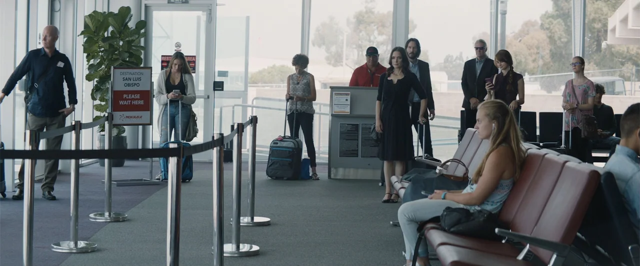 Destination Wedding (2018) movie still: wide — An airport boarding area with large windows and a check-in kiosk for passengers. Several people stand…