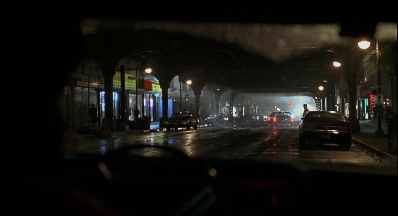 American Gangster (2007) movie still: establishing — A dark, wet city street is illuminated by overhead streetlamps and distant vehicle lights.…