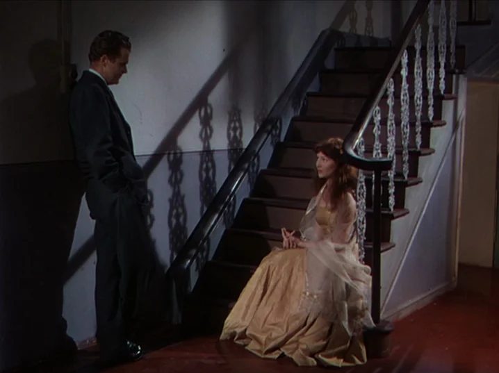 The River (1951) movie still: medium — A staircase interior with dark wood steps and patterned decorative metal railings. A man in a suit stands…