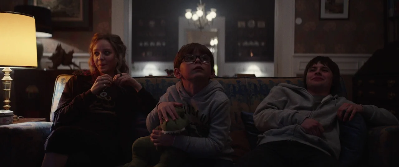 Colour Out of Space (2020) movie still: medium — A dimly lit room with patterned walls and a chandelier visible in the background doorway. Three…