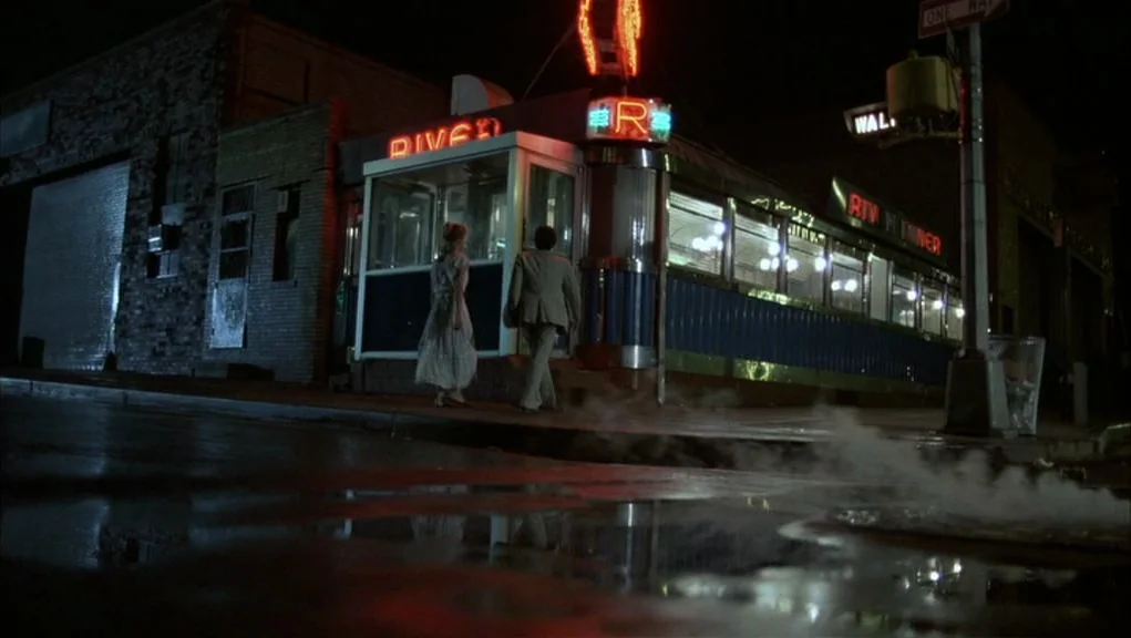 After Hours (1985) movie still: wide — A retro-style diner at night with glowing red neon signs on the dark street. Two people walk toward the…