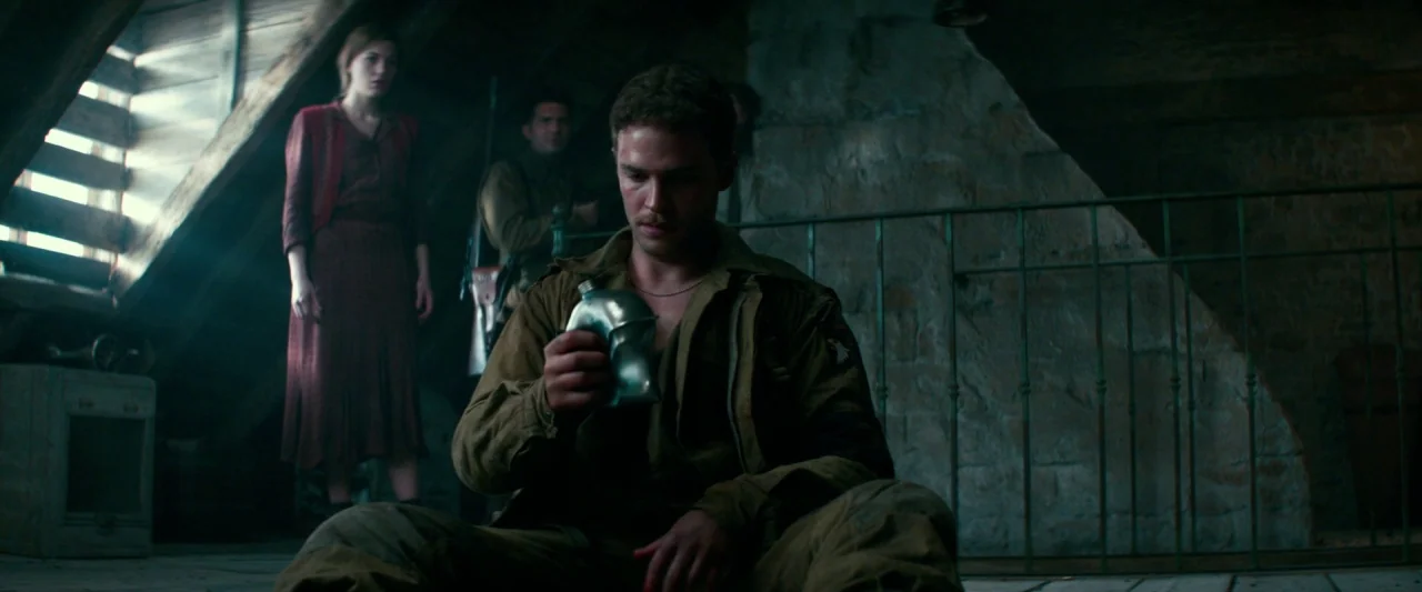 Overlord (2018) movie still: fullbody — A dimly lit attic features exposed wooden rafters and a rustic stone wall background. A young soldier sits…