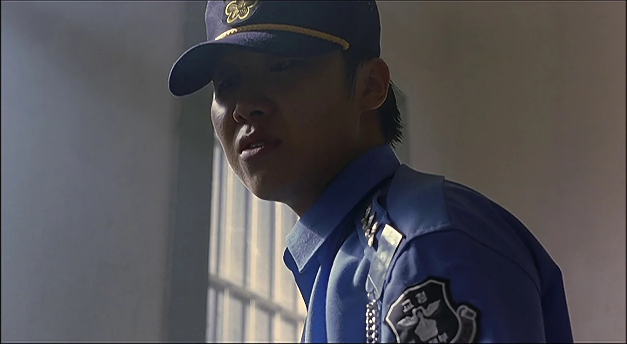 3-Iron (2004) movie still: medium — A man stands in an interior room with light filtering through a window. The subject wears a blue uniform with an…