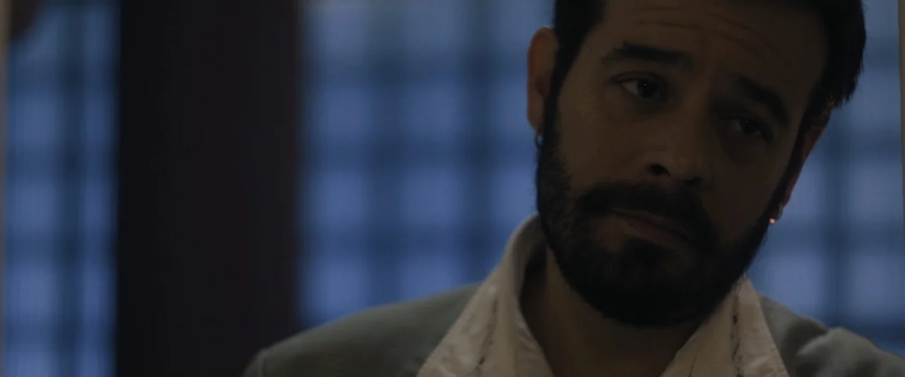 Sign Here (2017) movie still: closeup — A dimly lit room features a man positioned against a blurred dark blue background. The man with dark hair…