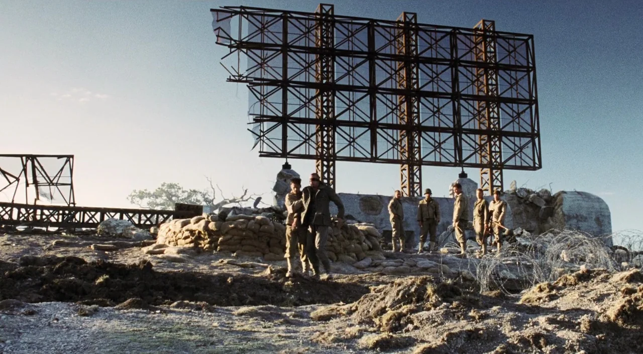 Saving Private Ryan (1998) movie still: wide — A war-torn landscape features a massive rusted metal billboard frame and sandbag fortifications. Two…