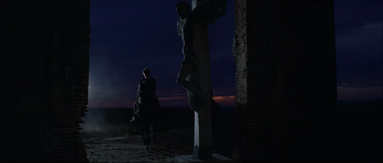 The Mercenary (1968) movie still: wide — A stone crucifix silhouette stands in the foreground of a ruined chapel archway. A man riding a horse…
