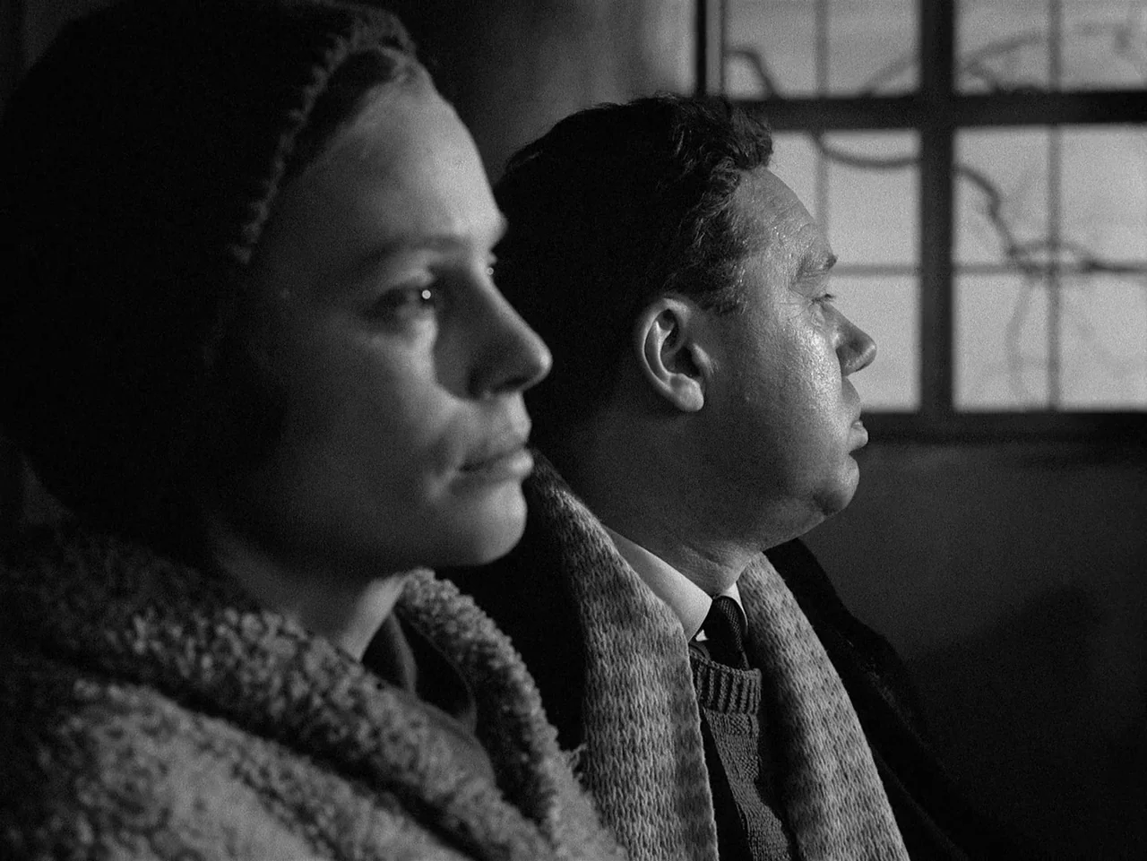 Winter Light (1963) movie still: medium — A man and woman sit next to each other in a dimly lit room. They look toward an off-screen source beside a…