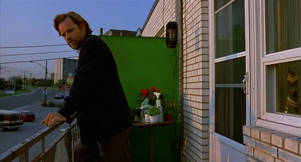 Exotica (1994) movie still: medium — A residential apartment balcony at sunset with a white brick wall and green screen. A man in a blazer leans…
