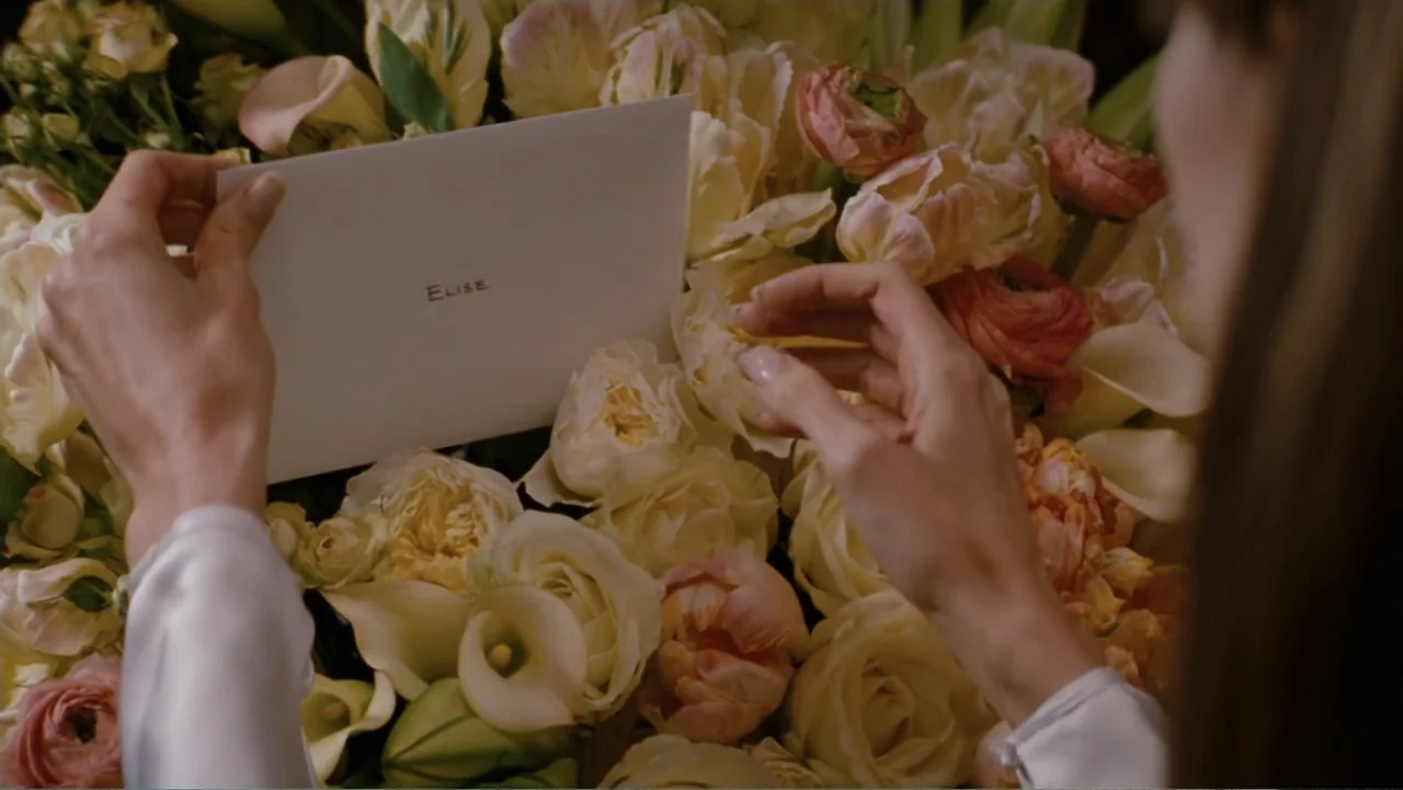The Tourist (2010) movie still: closeup — A close view shows a person holding a white envelope over a large flower arrangement. Pale cream and peach…
