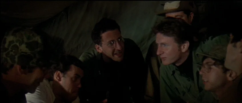 MASH (1970) movie still: medium — A group of men gather inside a dark military tent at night. They huddle together closely while engaged in a quiet…