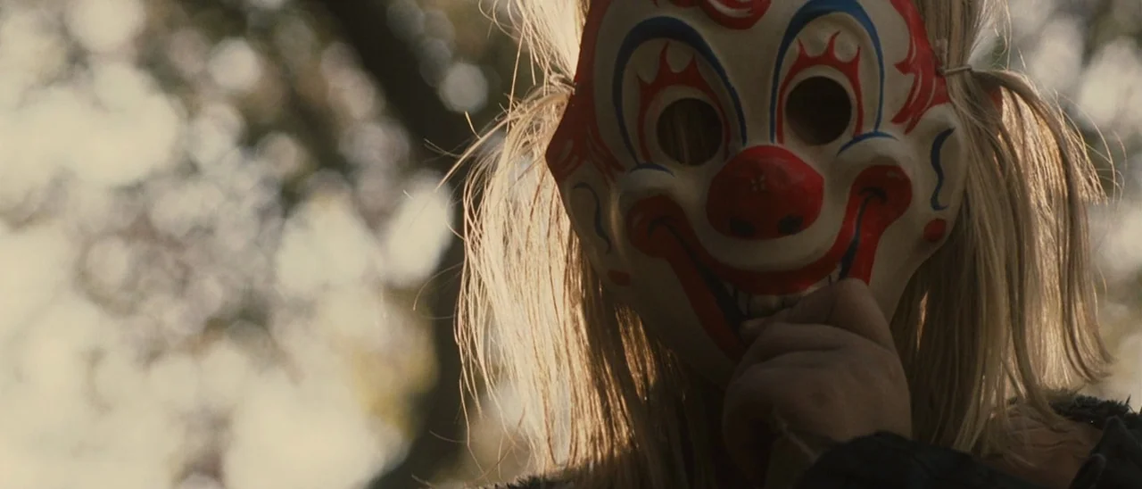 Halloween (2007) movie still: closeup — A sunlit outdoor area with blurred trees forming the background. A child wears a colorful plastic clown mask…
