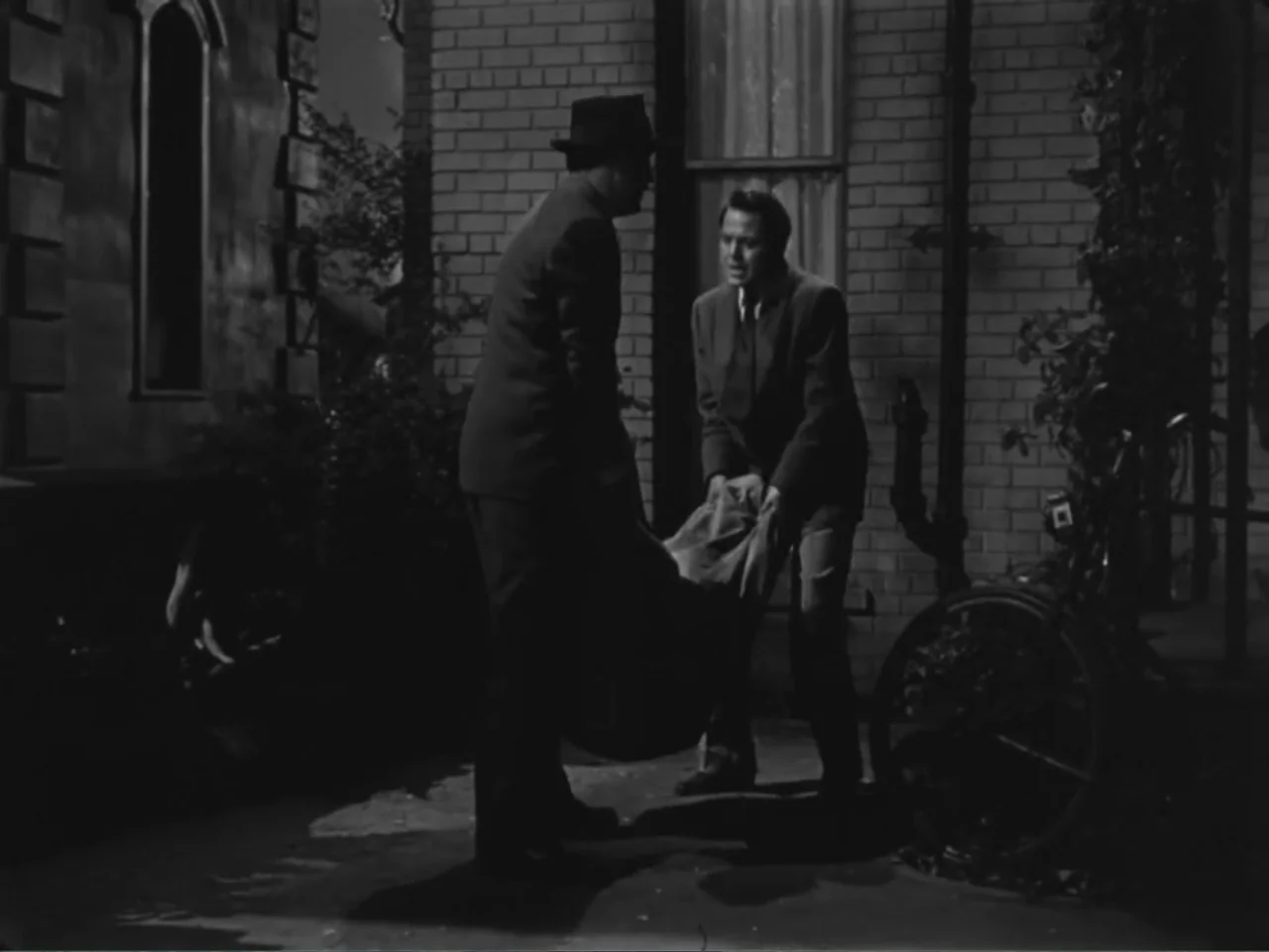 House by the River (1950) movie still: medium — A dark brick alleyway at night features a bicycle leaning against the wall. Two men in suits lift a…