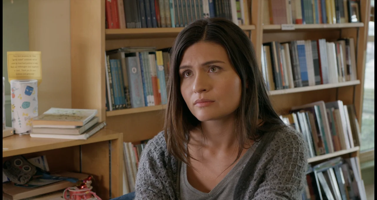 One True Loves (2023) movie still: medium — A woman sits in front of wooden shelves filled with various books. She looks slightly away from the…