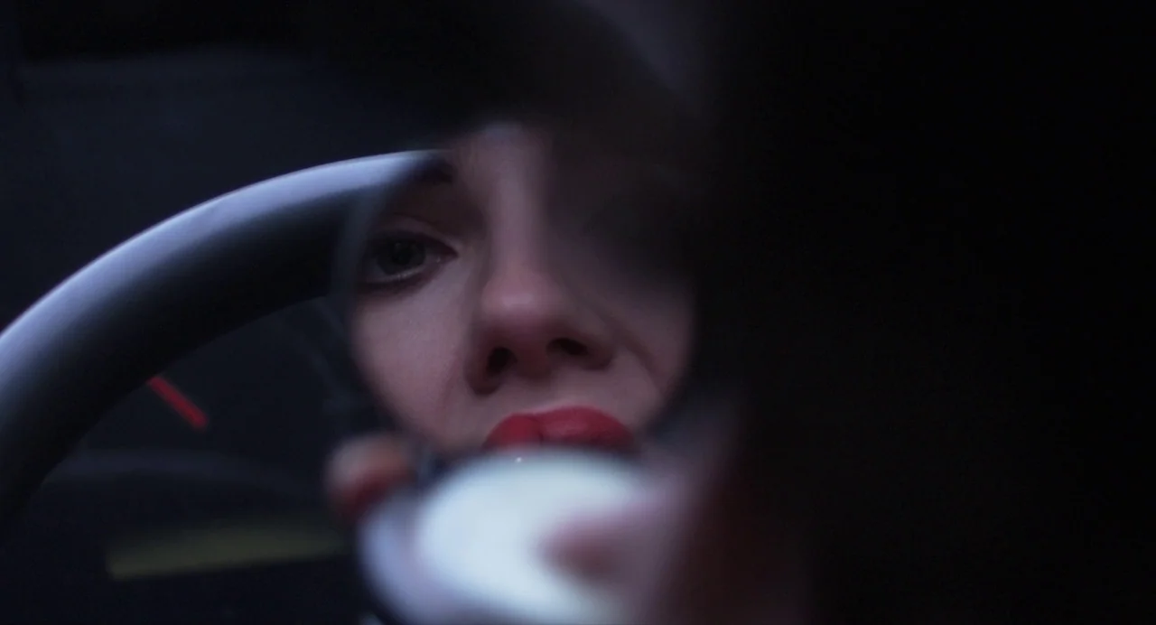 Under The Skin (2013) movie still: closeup — The interior of a dark car is illuminated by a small handheld mirror. A woman stares into the…