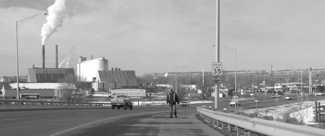 Nebraska (2013) movie still: wide — An industrial area with large factories and smoke stacks stands beside an open highway. A man walks down the…