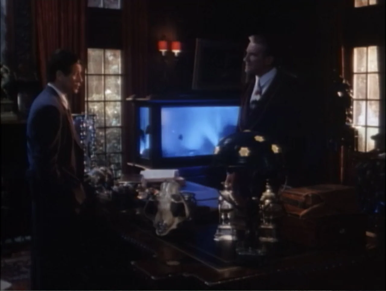 Cast a Deadly Spell (1991) movie still: medium — A dark, wood-paneled room features an illuminated aquarium resting on a large desk. Two men in…