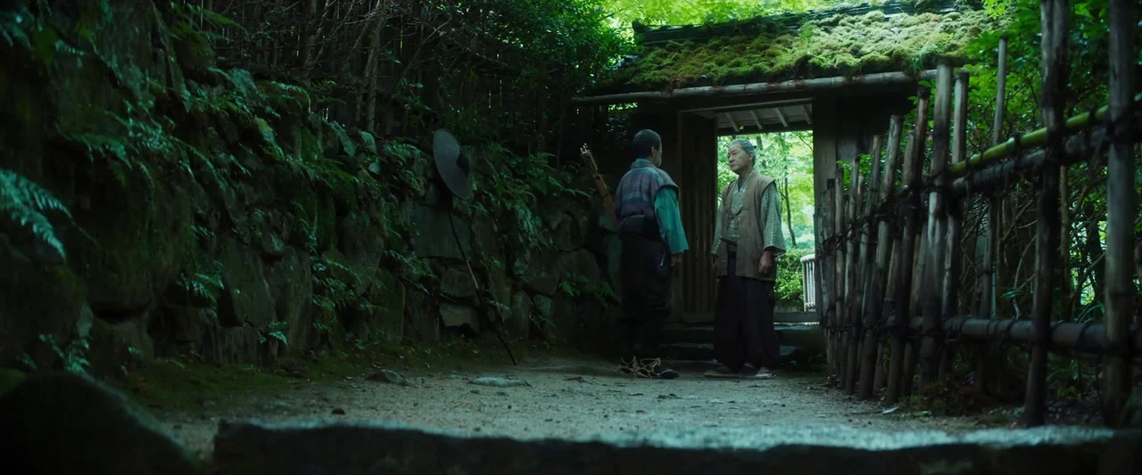 Kubi (2023) movie still: fullbody — A stone-walled path bordered by a bamboo fence leads to a gated structure. Two men stand in conversation before…