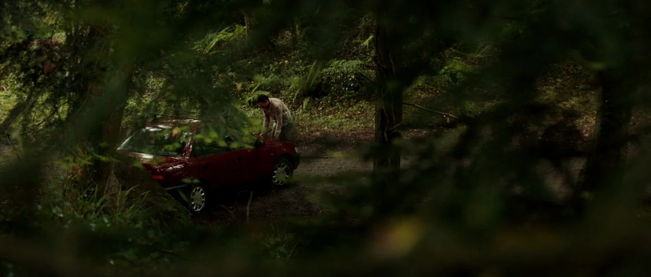 The Hallow (2015) movie still: wide — A dark red sedan is parked on a dirt trail surrounded by dense woodland. A man stands beside the driver side…