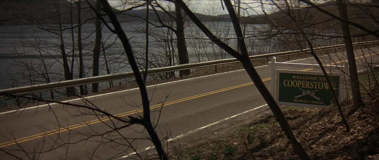 Cobb (1994) movie still: wide — A rural asphalt road curves past a lake, bordered by bare, leafless trees. A green Welcome to Cooperstown sign…