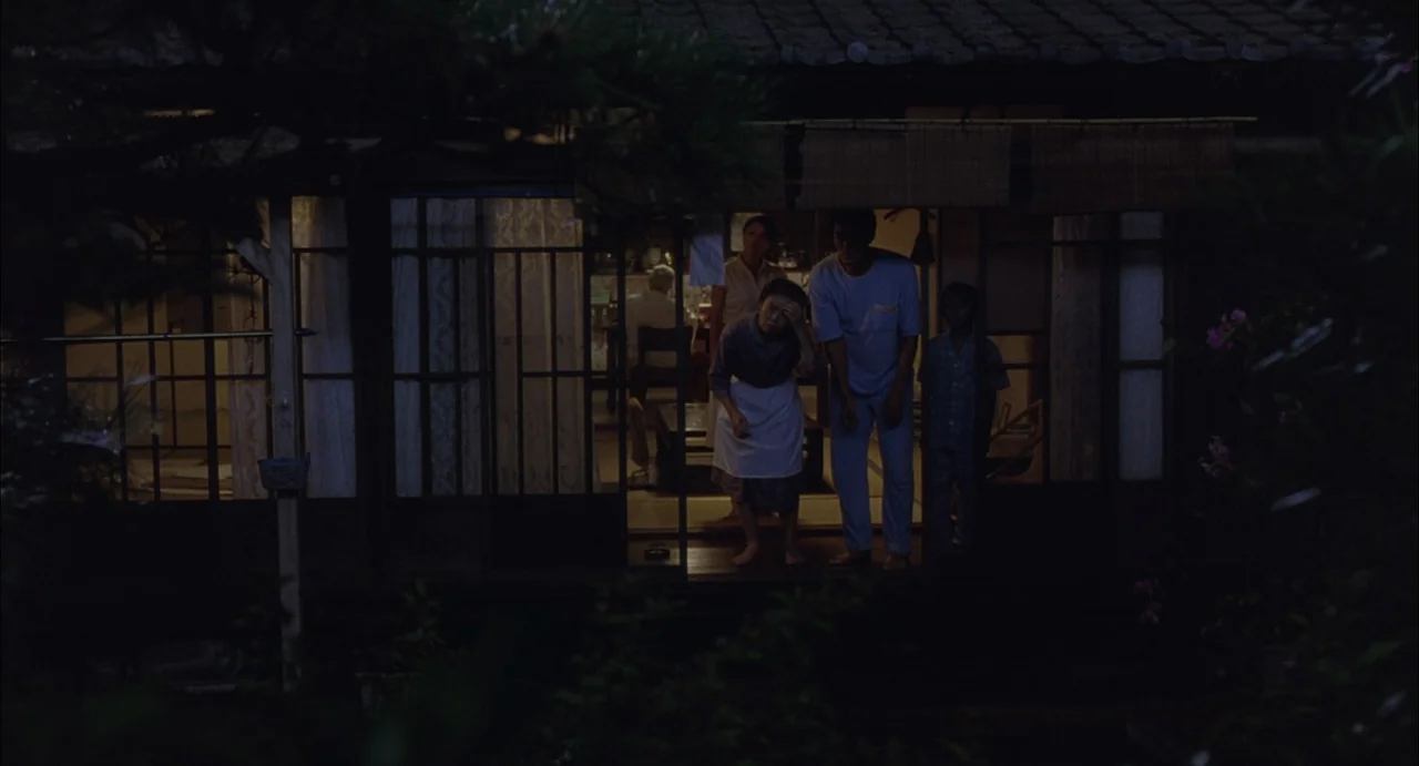 Still Walking (2008) movie still: fullbody — A group of people stand on a Japanese wooden porch at night. They look out from a dimly lit interior…