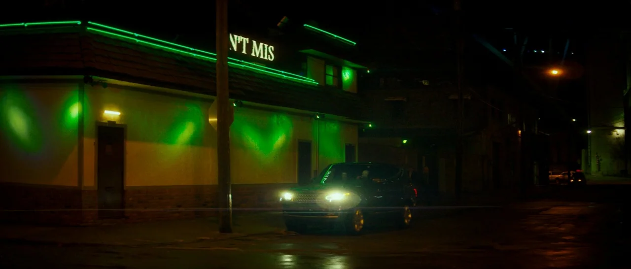 Dog Eat Dog (2016) movie still: wide — A dark city building features a prominent green neon sign reading N'T MIS. A dark SUV is parked at the curb…