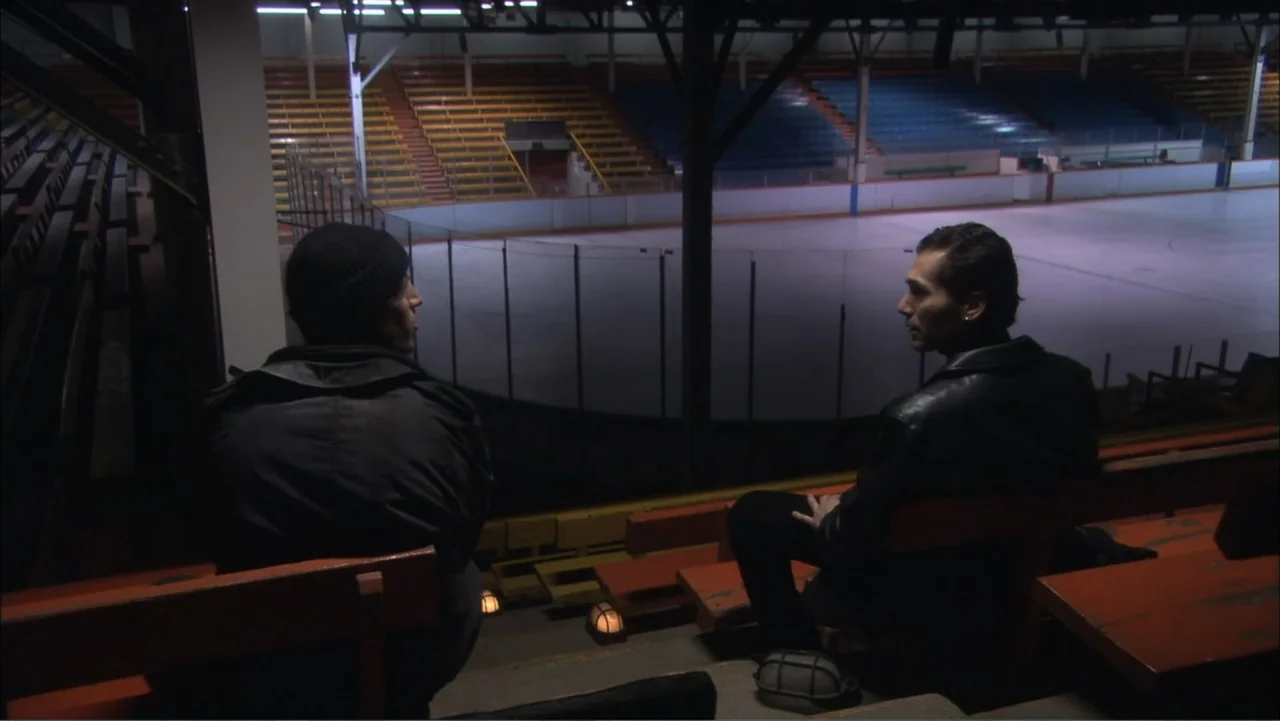 O código oculto (2025) movie still: medium — Empty orange wooden bleachers overlook a large indoor ice rink floor in a dim stadium. Two men sit…
