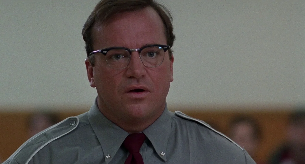 Touch (1997) movie still: closeup — A man wearing a grey uniform stands in front of a neutral-toned wall inside a building. He is looking forward…