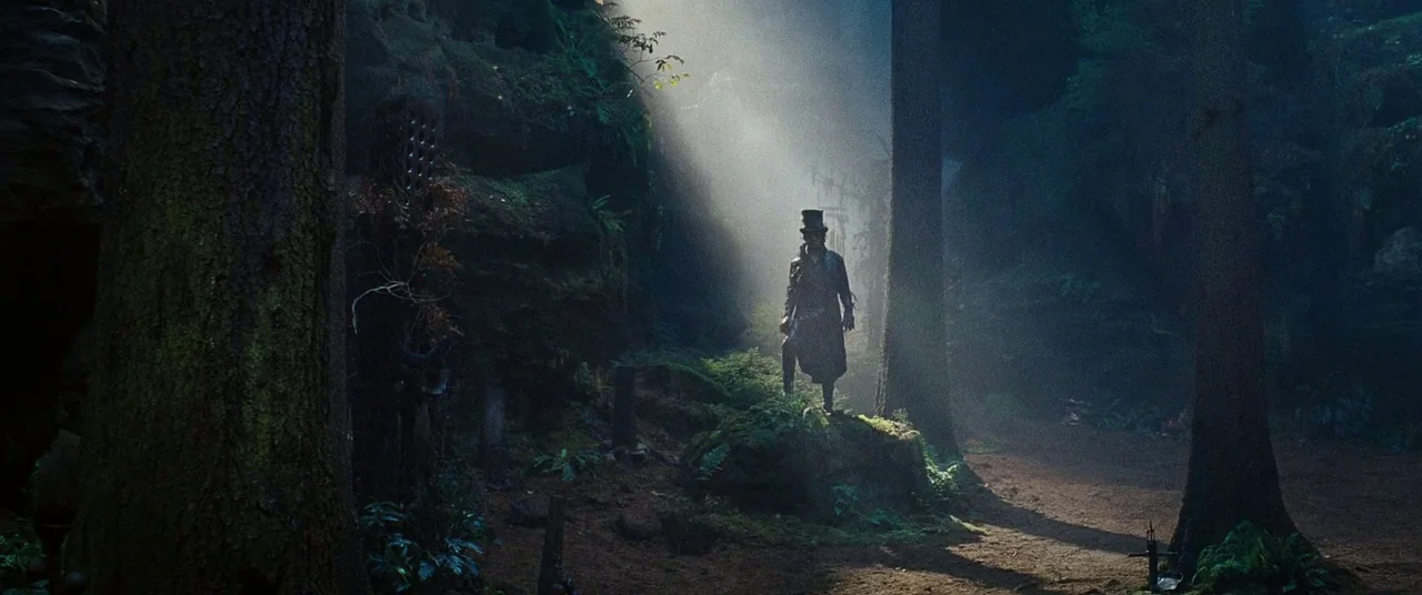 Cloud Atlas (2012) movie still: wide — A dark, atmospheric woodland with towering evergreens and mossy, rocky outcrops. A man in a top hat stands…