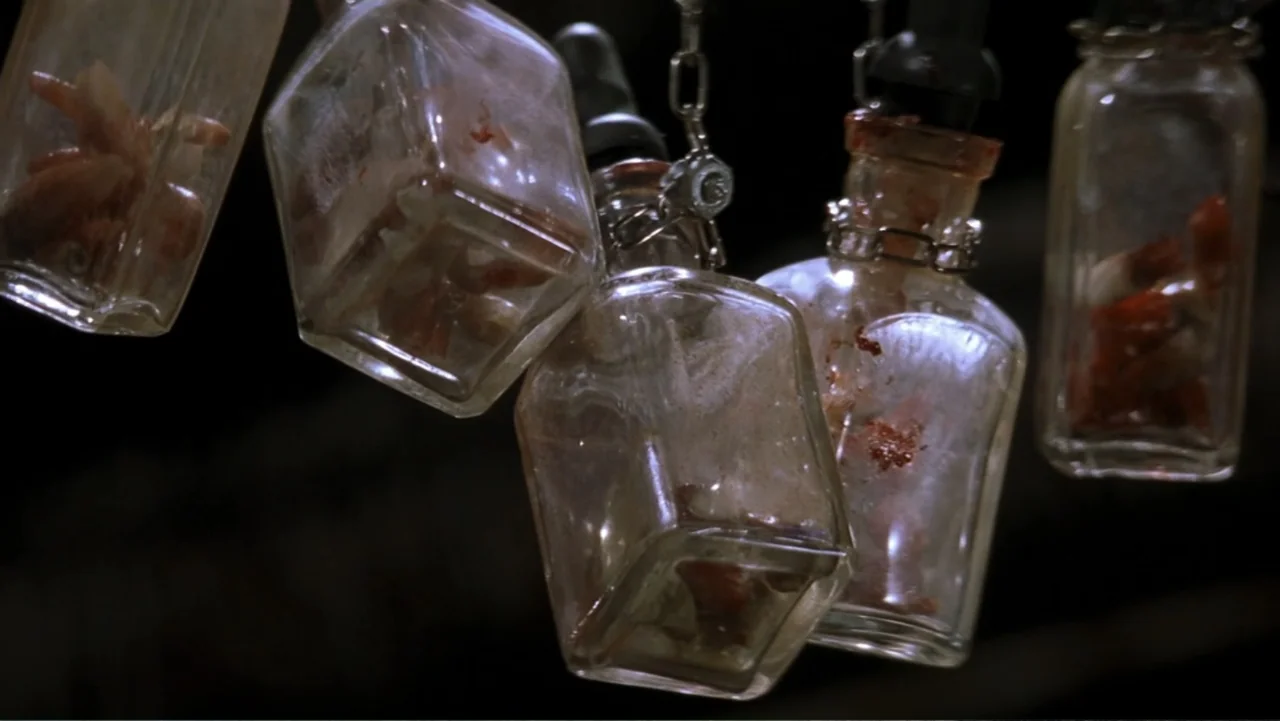 Ghosts of Mars (2001) movie still: closeup — Several clear glass medical vials hang from metal chains against a dark backdrop. Each container holds…