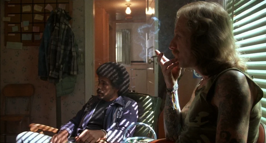 Drugstore Cowboy (1989) movie still: medium — A cluttered apartment space is illuminated by light streaming through striped window blinds. A…