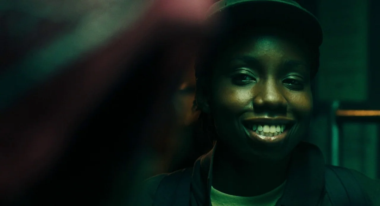 Pariah (2011) movie still: closeup — A dimly lit indoor space with a dark, green-toned lighting scheme. A young woman wears a cap and smiles while…