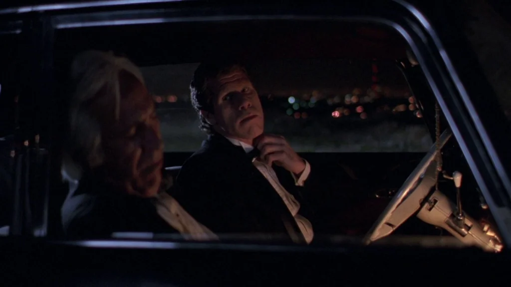 Cronos (1993) movie still: medium — The interior of a parked car at night overlooking a distant city. A man in a tuxedo adjusts his neckwear while…
