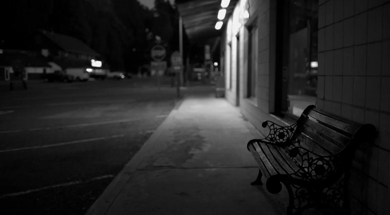 Blue Jay (2016) movie still: wide — An empty dark bench rests on a concrete sidewalk beside a tiled storefront building. The background shows a…