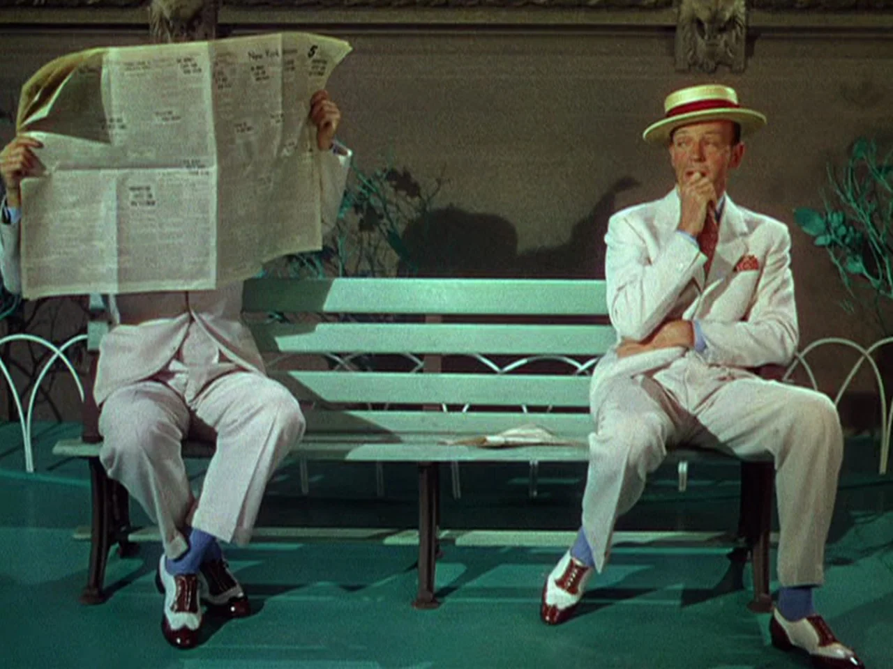Ziegfeld Follies (1945) movie still: fullbody — Two men in white suits sit on a pale green wooden bench. One man holds a large newspaper obscuring…
