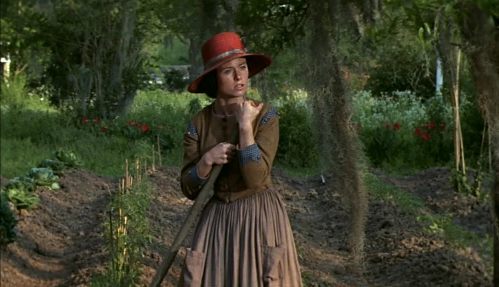 The Beguiled (1971) movie still: medium — A rural vegetable garden with rows of tilled earth and leafy green crops. A woman in a brown period dress…