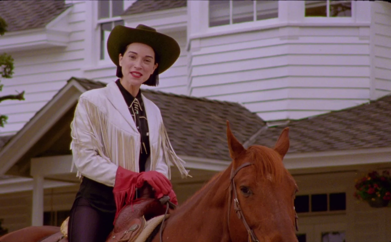 The Nowhere Inn (2020) movie still: medium — A woman in western-style clothing sits on a horse in front of a white house. The house features classic…