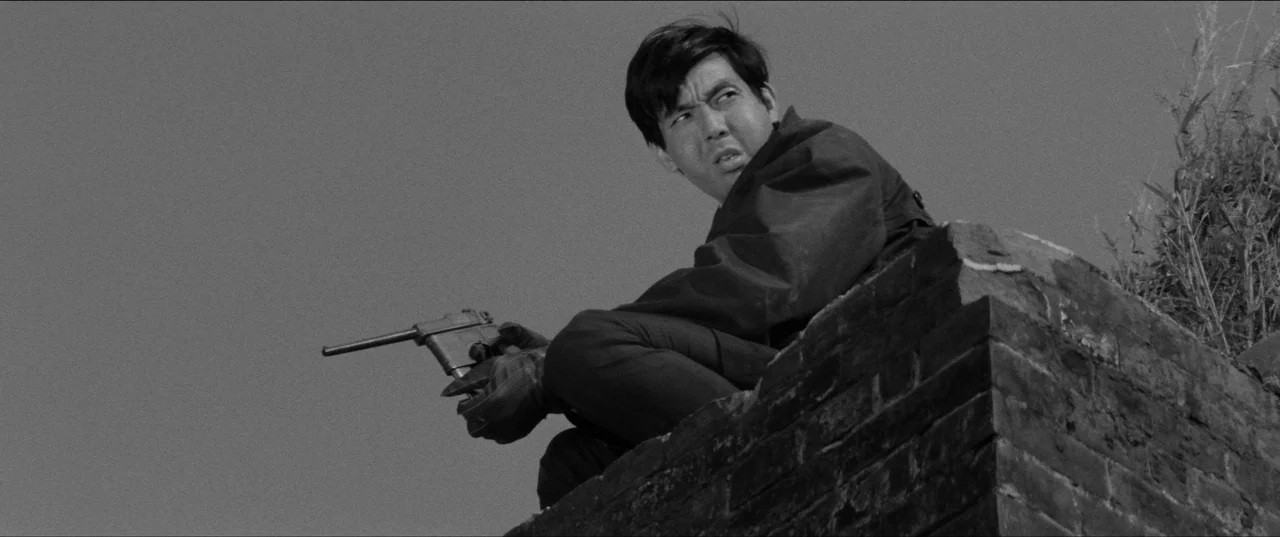 Branded To Kill (1967) movie still: medium — A man crouches on the edge of a brick structure against a flat sky. He holds a pistol while looking…