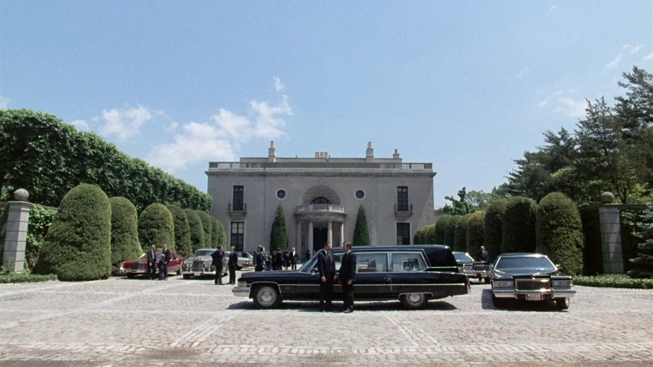 The Sentinel (1977) movie still: wide — A stone mansion with a large semicircular driveway stands beneath a clear blue sky. Two men in black suits…