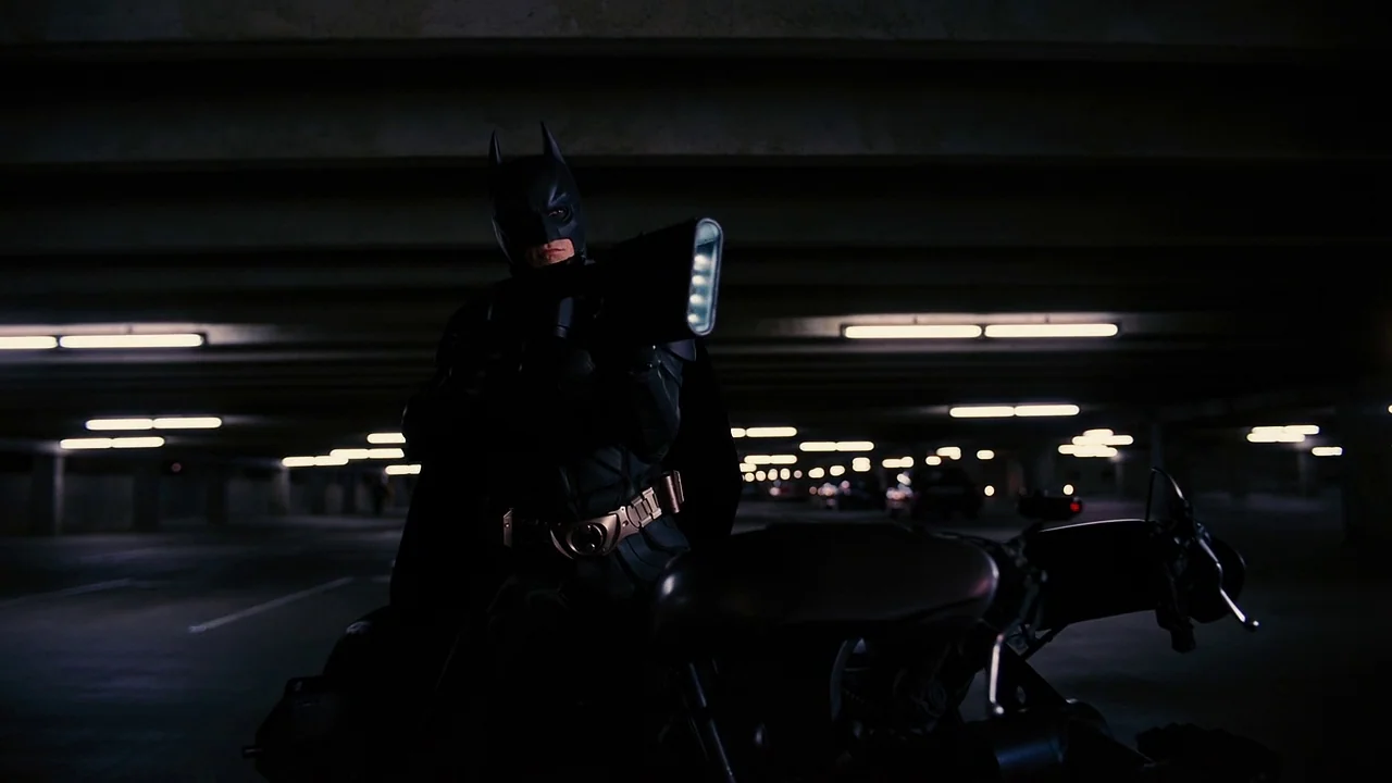 The Dark Knight Rises (2012) movie still: medium — A dark, empty concrete parking garage features rows of repetitive overhead fluorescent lighting…