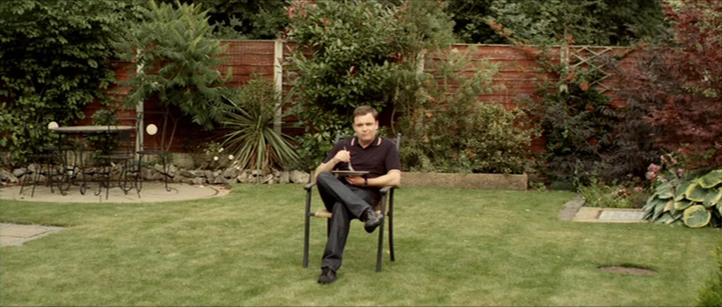 Kill List movie still: fullbody — A quiet residential backyard features a manicured lawn enclosed by a wooden privacy fence. A man sits alone in the…