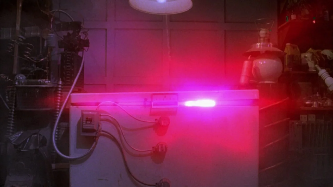 Frankenhooker (1990) movie still: medium — A cluttered workshop space contains a white chest freezer modified with exposed electrical wiring. The…
