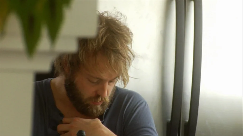 Humpday (2009) movie still: medium — A man sits indoors with a bright light source visible off to his right. He gazes downward toward his own chest…