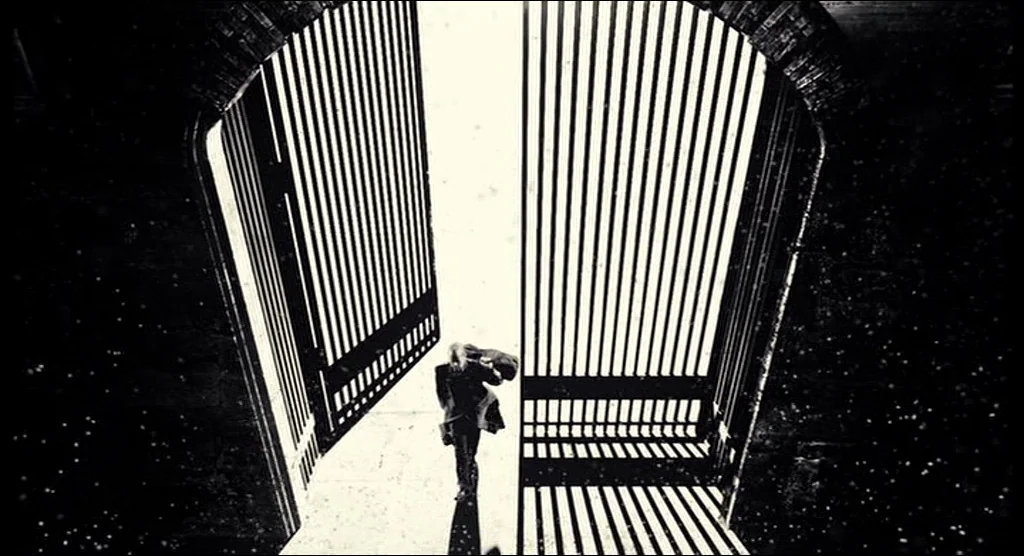 Sin City (2005) movie still: wide — A high-angle view looks down at a figure passing through massive metal gates. Snow or dust particles fall…