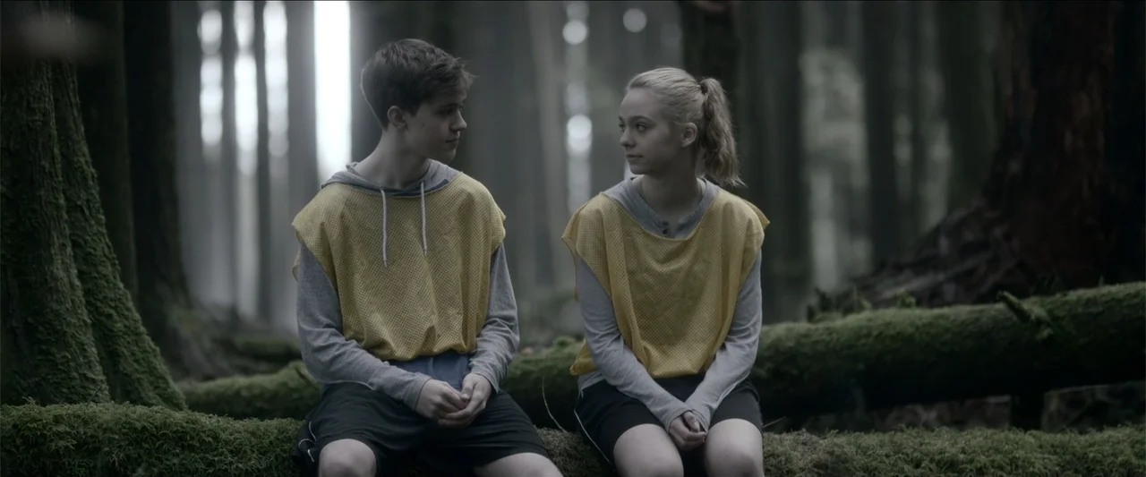 The Hollow Child (2018) movie still: medium — A dense forest with mossy logs and tall coniferous trees forms the environment. Two teenagers wearing…