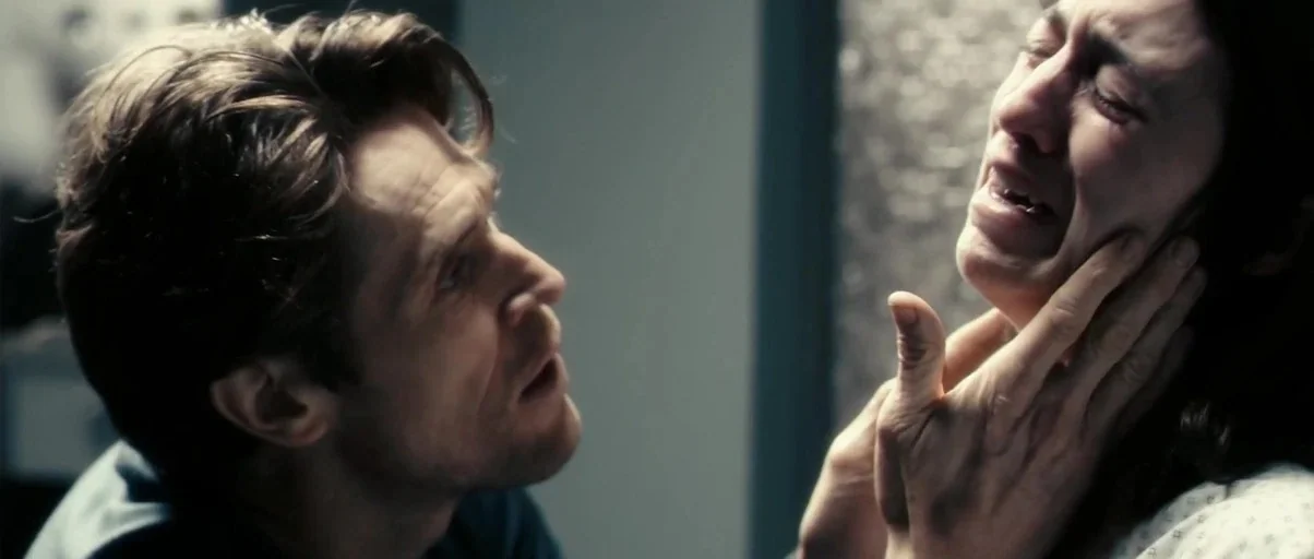 Antichrist (2009) movie still: closeup — A dimly lit room with muted neutral tones and blurred medical surroundings. A man leans in closely,…