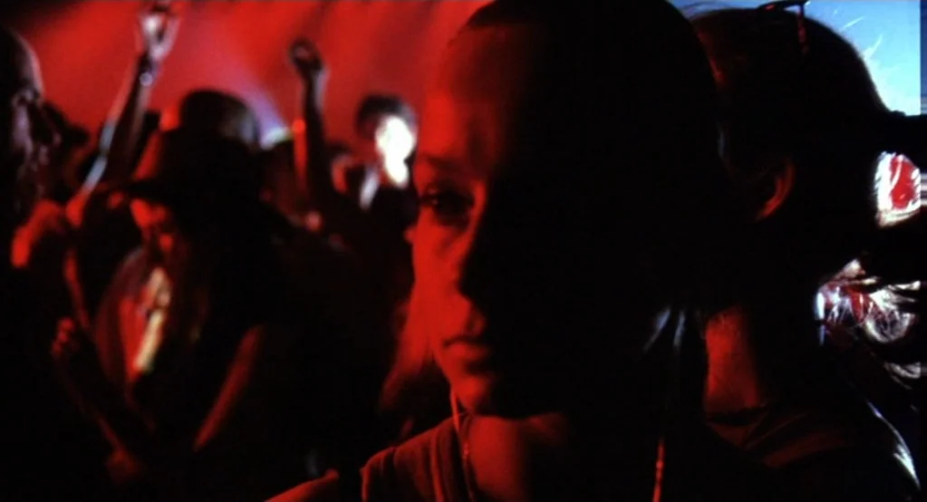 Morvern Callar (2002) movie still: closeup — A crowded, dimly lit nightclub interior with people dancing in the background. A young woman stands in…