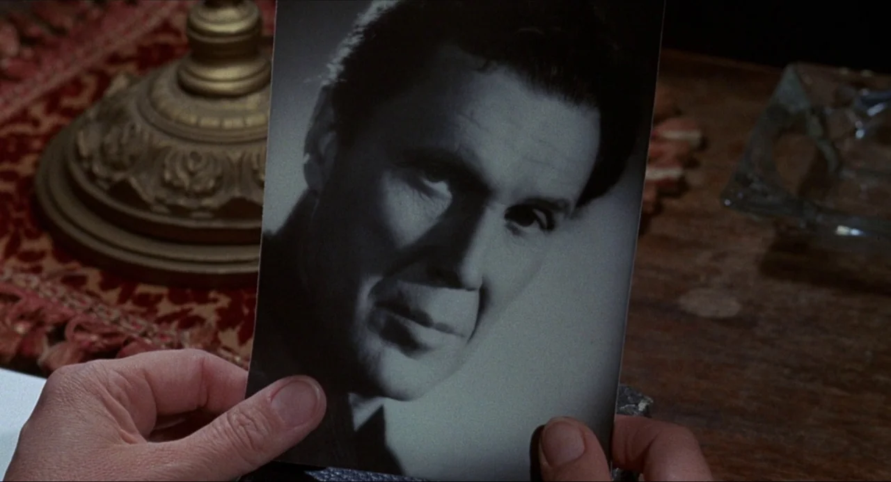 What’s the Matter with Helen? (1971) movie still: closeup — A wooden table surface sits beneath a photograph showing a man's portrait. Two hands…