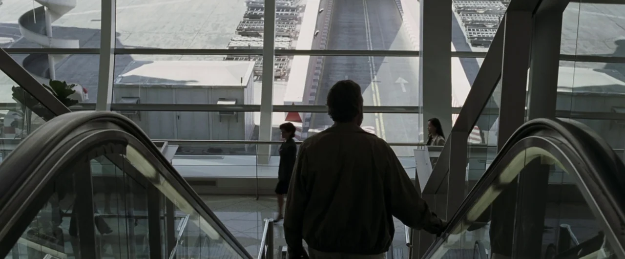 Syriana (2005) movie still: wide — A spacious airport terminal with large floor-to-ceiling glass panels overlooking a tarmac road. A man in a tan…