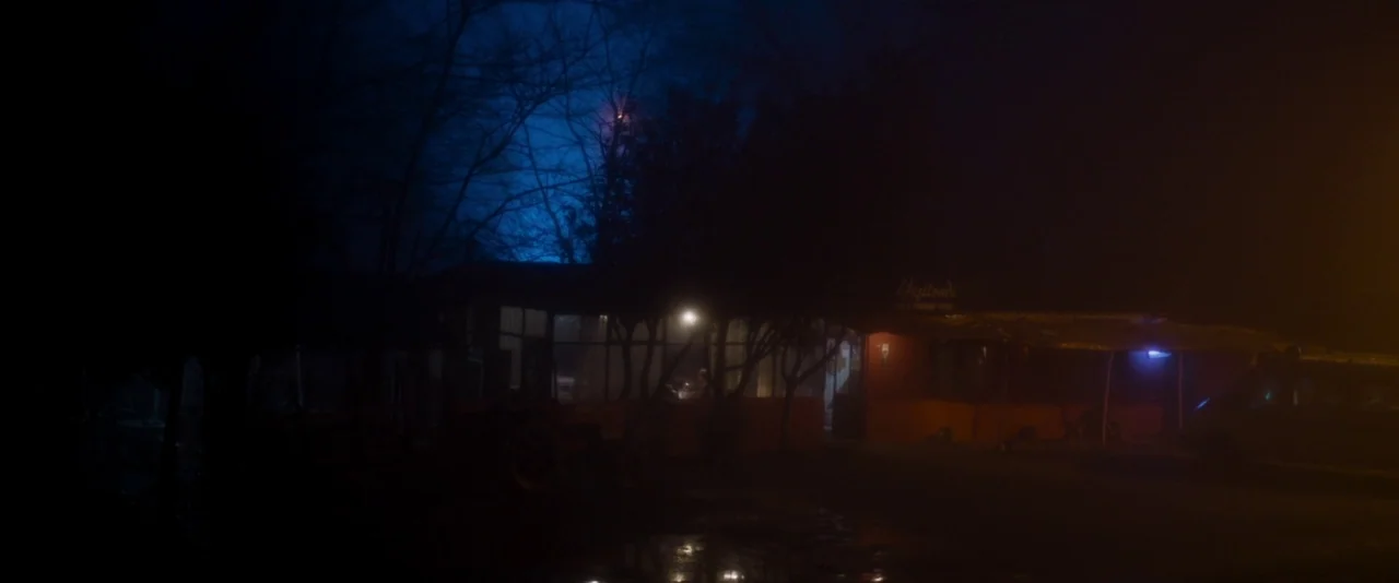 Baskin (2015) movie still: establishing — A low-slung restaurant building sits at night under a deep blue sky. Dark trees frame the structure which…