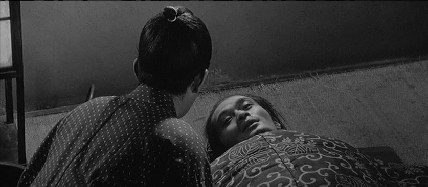 Red Beard (1965) movie still: medium — A dark, spare interior space with a woven tatami floor and simple wall. A person in a patterned kimono stares…